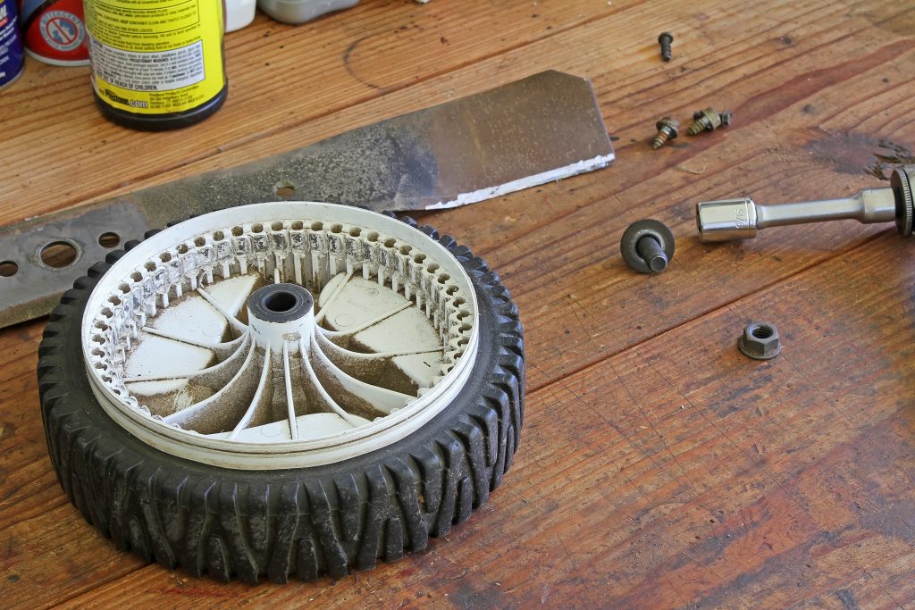 Closeup of plastic lawn mower drive-wheel with drive gear teeth completely destroyed. Sharpened lawn mower blade in background with nuts bolts tools all on wooden workbench.